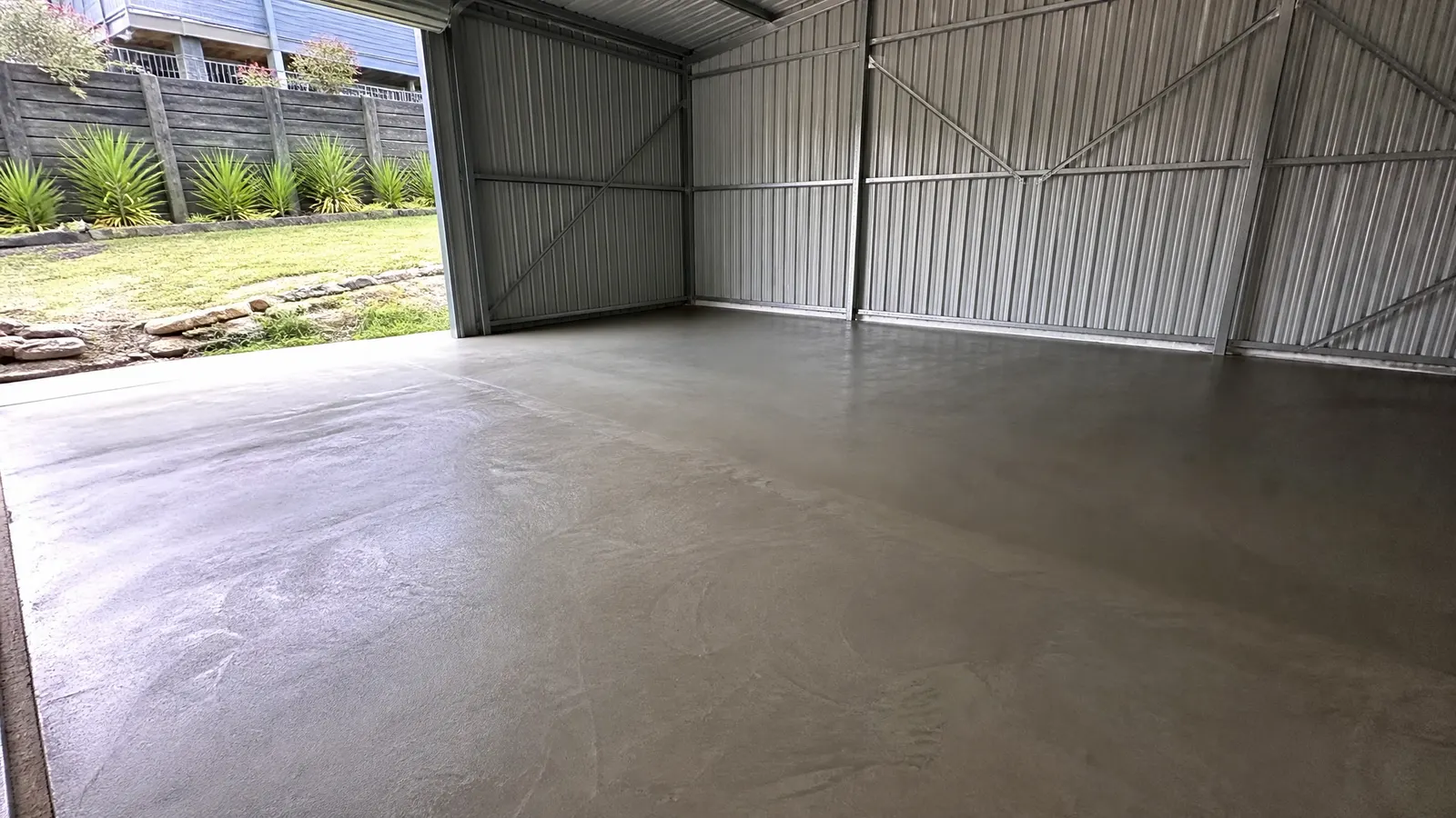 Large concrete workshop slab freshly finished with trowelled surface next to established garden and fence on Gold Coast property