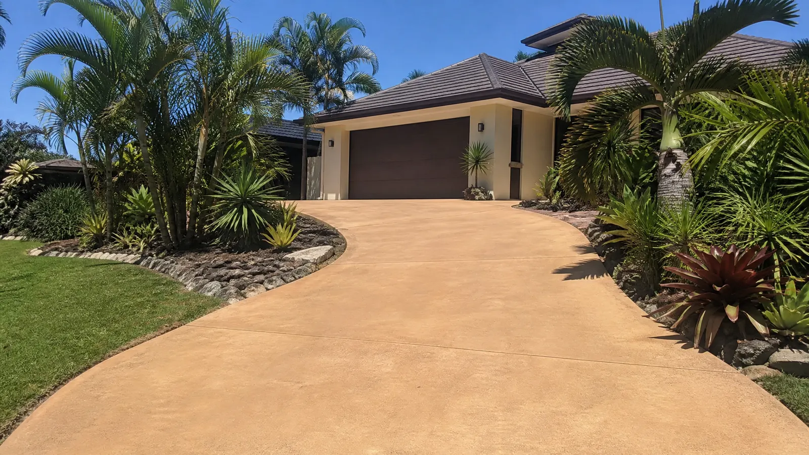 Smooth coloured concrete driveway in warm sandstone tone leading to a modern Gold Coast home with landscaped garden borders